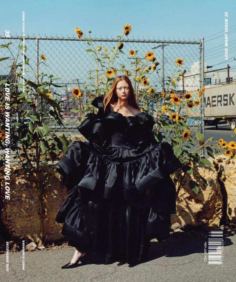 A fashionable woman in a large black dress poses outdoors near sunflowers and a chain-link fence, with a shipping container in the background.