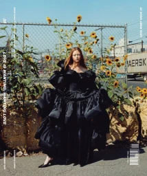 Model standing outdoors wearing an oversized black ruffled gown in front of a chain-link fence with sunflowers.