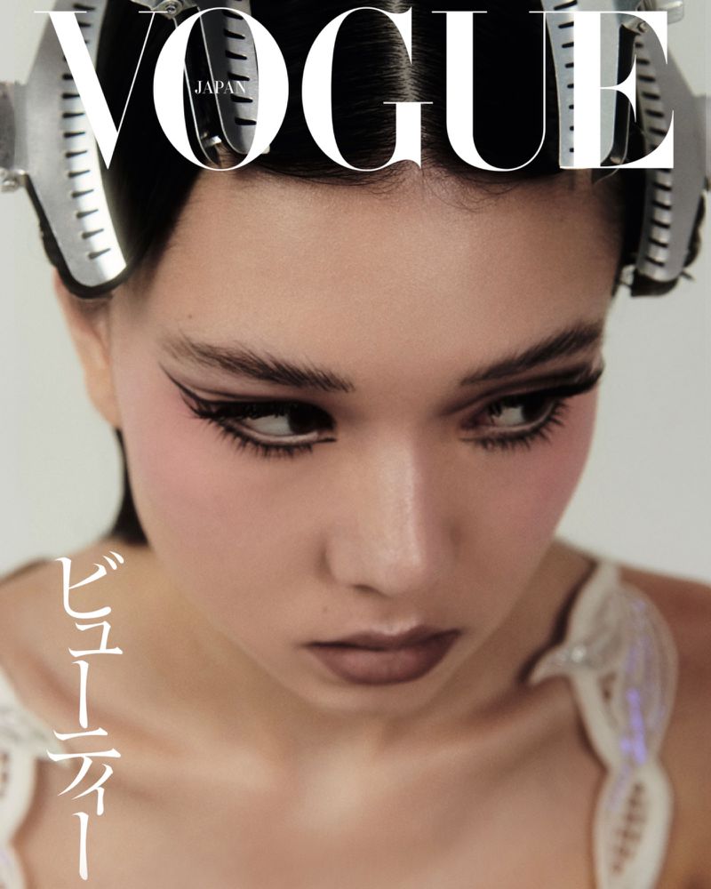 Close-up of a woman's face on a Vogue Japan cover, featuring dramatic winged eyeliner, pink blush, hair curlers, and a white background.