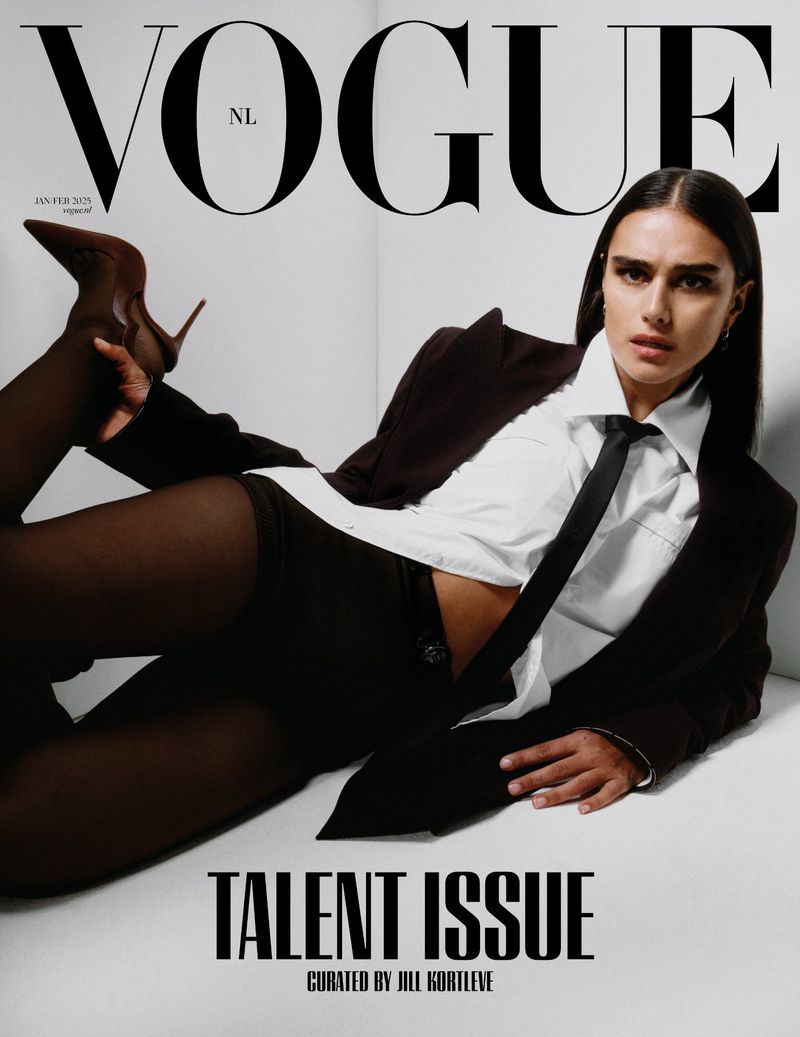 Vogue NL cover featuring a woman in a blazer and tights, reclining on a white surface, with the magazine's title in large black letters.