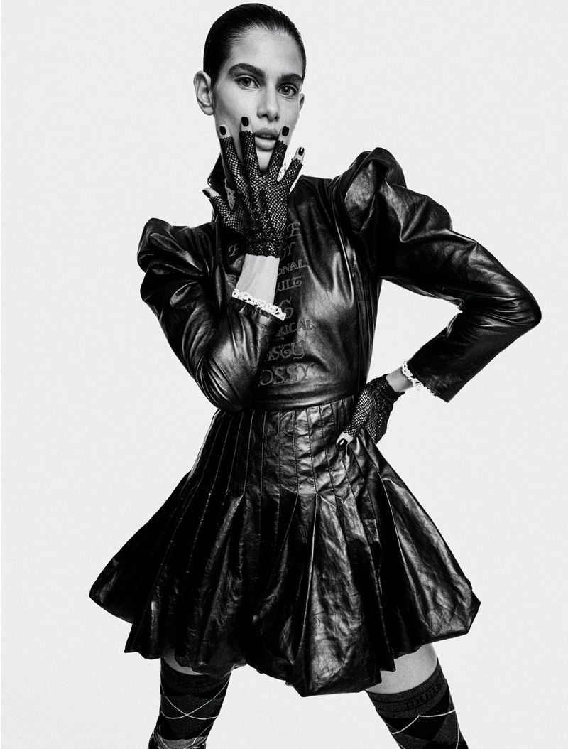 A woman poses in a black leather dress, gloves, and argyle stockings against a white backdrop for a fashion editorial shoot.