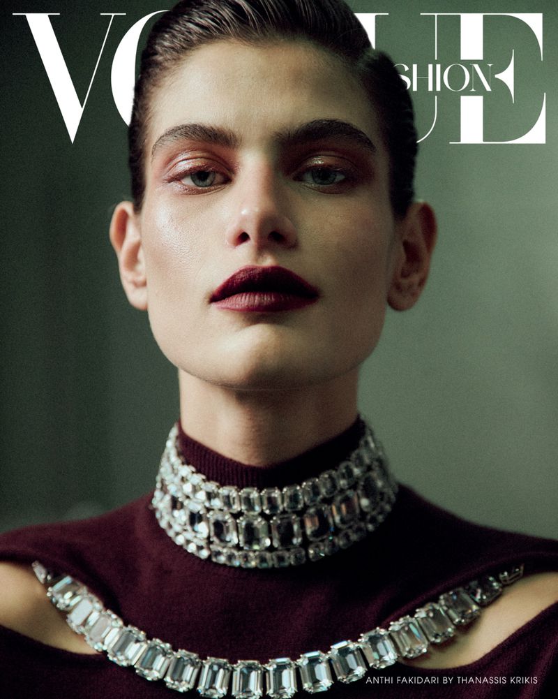 Fashion editorial shot of a woman with dark hair and makeup wearing jeweled necklaces in front of a blurred studio backdrop.