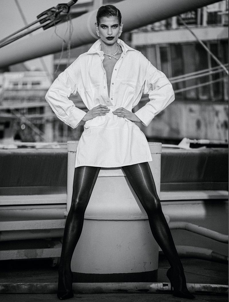 A monochrome portrait of a female model in a white shirt dress, black tights, and high heels, posing against an industrial backdrop.