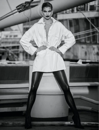 A monochrome portrait of a female model in a white shirt dress, black tights, and high heels, posing against an industrial backdrop.