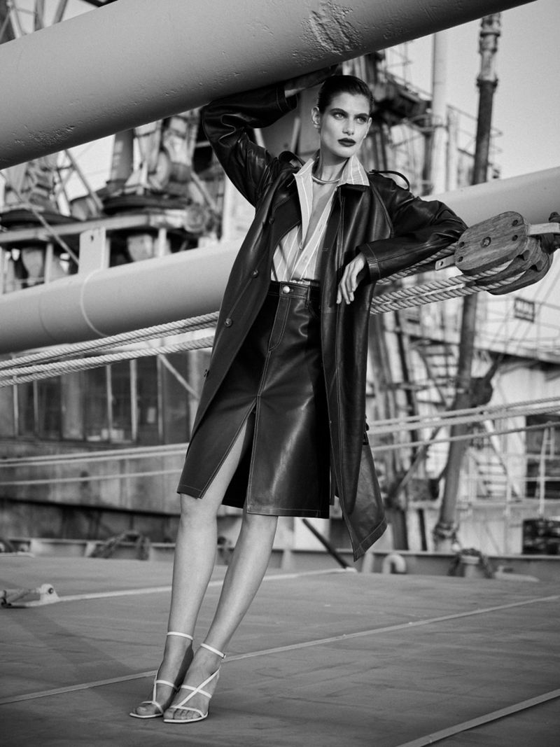 Monochrome fashion shot of an adult female model wearing a leather skirt, coat, and strappy heels, leaning against a pipe structure outdoors.