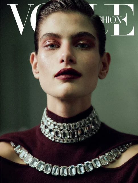 Close-up of a fashion magazine cover featuring a model with slicked-back hair, burgundy makeup, and an elaborate jeweled collar necklace.