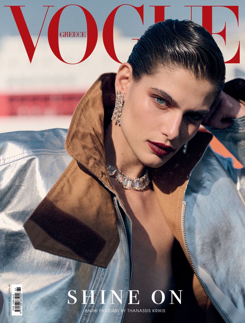 Vogue Greece cover featuring a model with slicked-back hair, wearing a metallic jacket and jewelry, with a blurry background.
