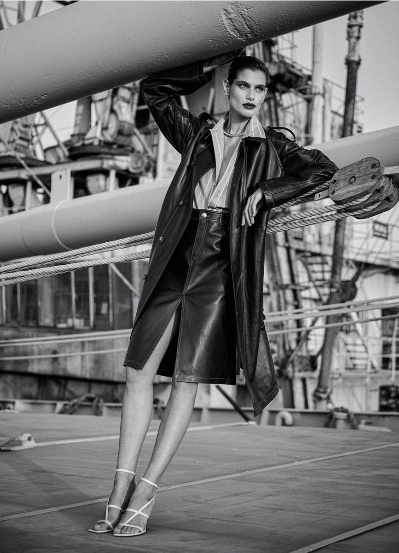 Elegant adult woman in a black leather trench coat and skirt poses on an industrial structure with heavy cables and a textured gray background.