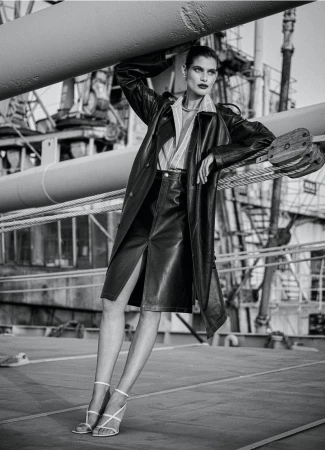 Elegant adult woman in a black leather trench coat and skirt poses on an industrial structure with heavy cables and a textured gray background.