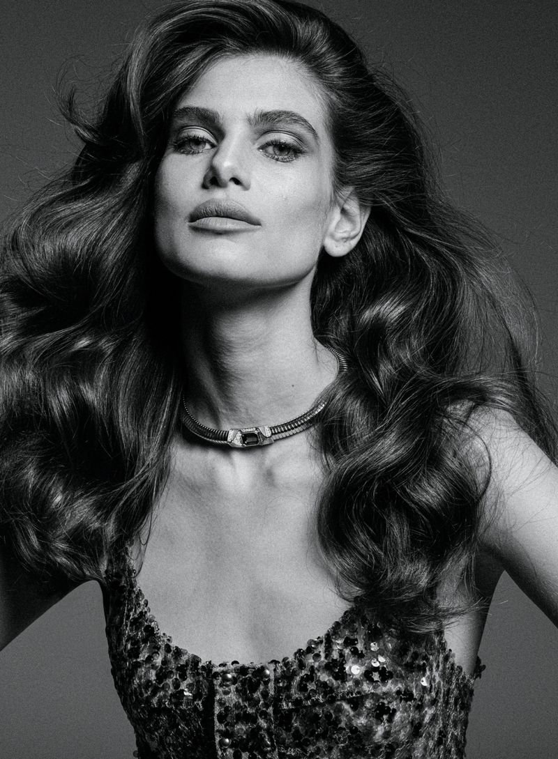 Monochrome close-up portrait of an adult woman with long wavy hair, a choker necklace, and a sequined top against a grey background.