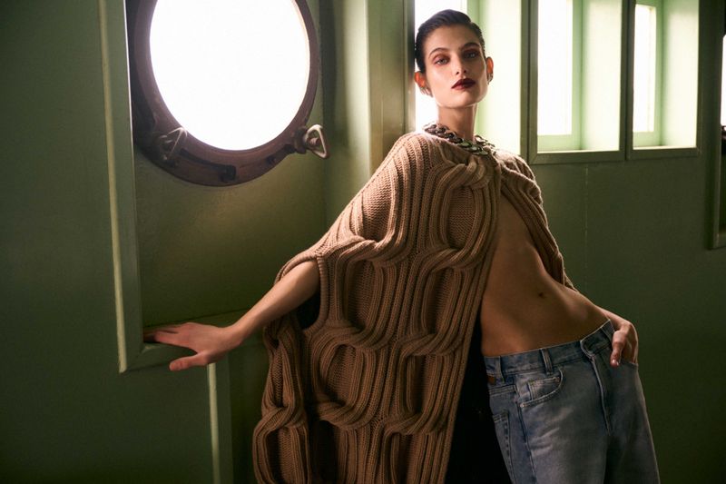 Fashionable adult woman in a tan knit poncho and jeans, posing near a porthole window in an indoor setting with soft studio lighting.