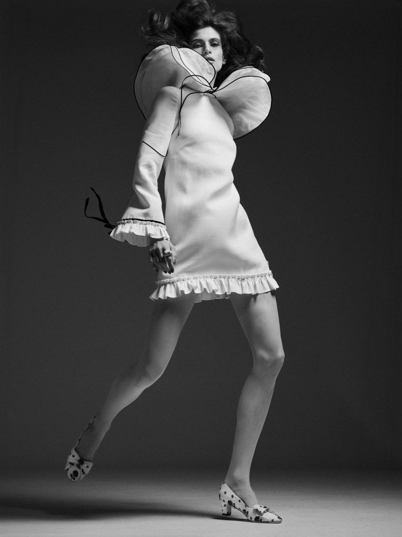 Fashionable woman in a white ruffle-trim dress strikes a pose in a studio setting, showcasing a blend of style and high fashion.
