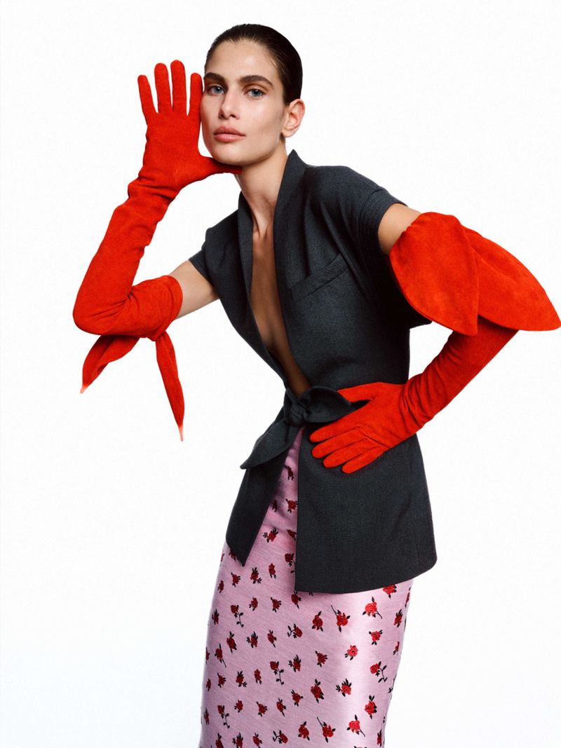 Fashion shot of a woman in a gray blazer, floral skirt, and long red gloves, posing on a white background in a studio setting.