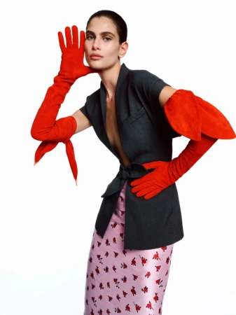 Fashion shot of a woman in a gray blazer, floral skirt, and long red gloves, posing on a white background in a studio setting.