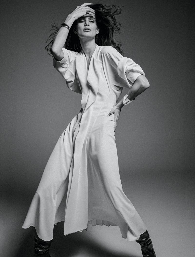 Monochrome fashion shot of an adult woman in a long white coat dress and dark boots, posing in a photo studio with hand on her head.