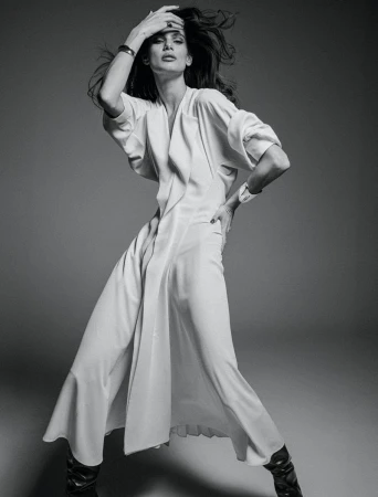 Monochrome fashion shot of an adult woman in a long white coat dress and dark boots, posing in a photo studio with hand on her head.