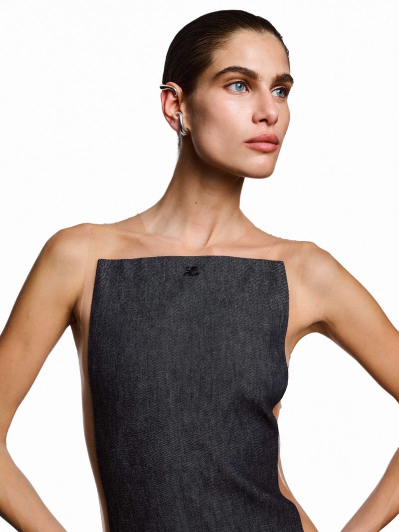 Portrait of a woman in a studio with a dark gray, rectangular garment, silver ear jewelry, slicked-back hair, and a white background.