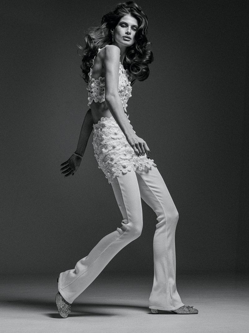 An adult female fashion model wearing a white flower-applique top and white flared pants poses in a studio with dramatic lighting.