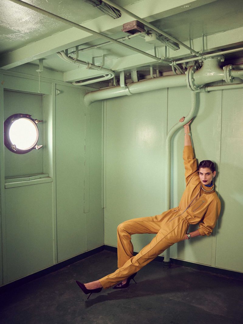 An adult female fashion model in a yellow jumpsuit poses while holding onto pipes inside a mint green painted room with a porthole.