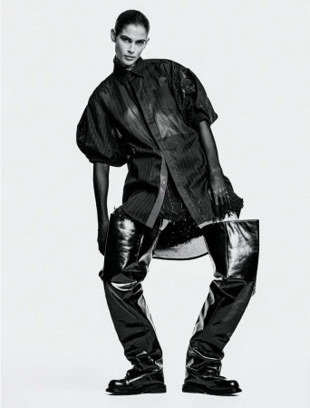 Model Anthi Fakidari poses in an oversized dark shirt and dramatic glossy black thigh-high boots against a seamless white studio background.