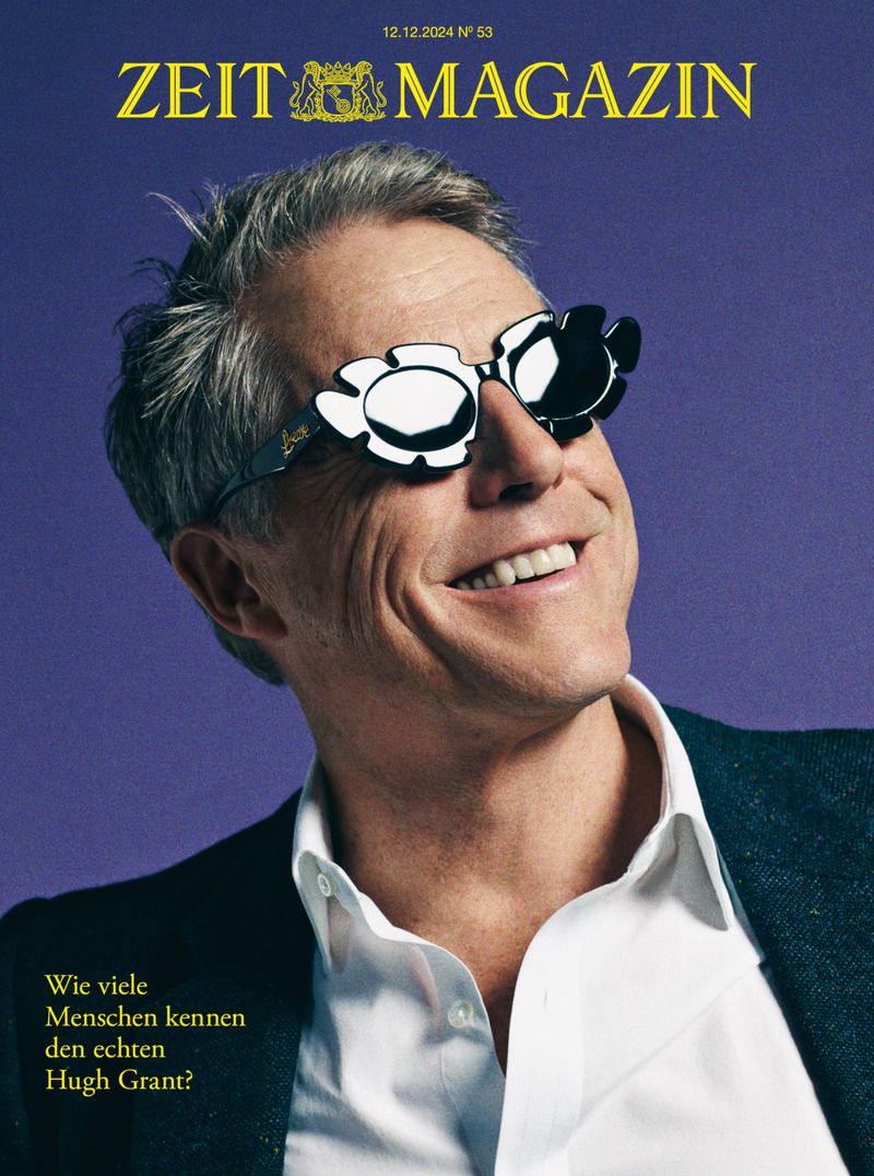 Magazine cover featuring a smiling man with flower-shaped sunglasses in a suit and white shirt against a purple backdrop.