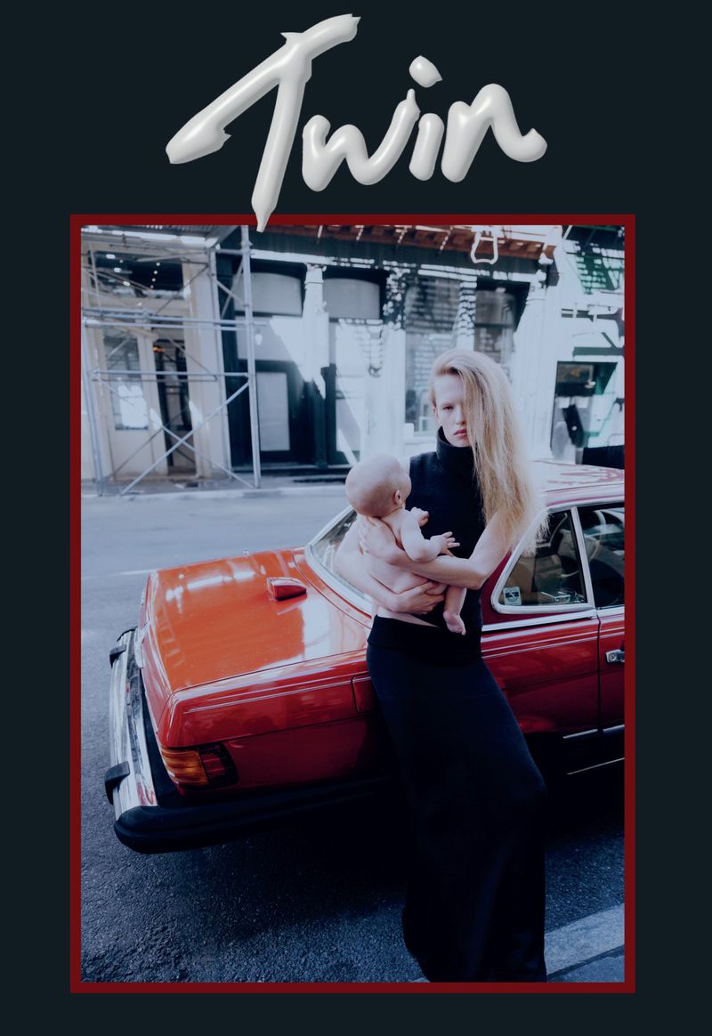 Fashion editorial shot of a woman in a black dress holding a baby next to a red car in an urban setting for Twin magazine.