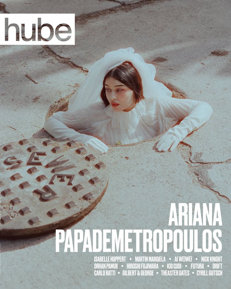 Artistic fashion portrait of a young woman emerging from a concrete hole cover, wearing white dress, white gloves, pale skin and makeup.