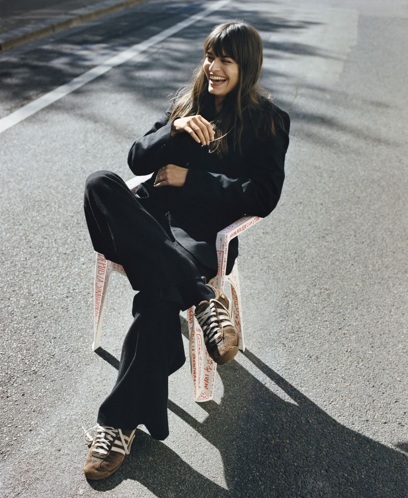 A woman in a black suit and brown sneakers sits outside on a white chair and laughs in an eye-level medium shot on a sunny day.