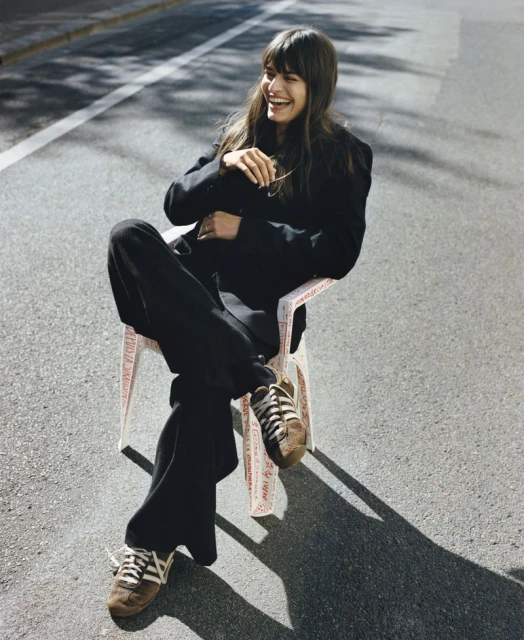 A woman in a black suit and brown sneakers sits outside on a white chair and laughs in an eye-level medium shot on a sunny day.