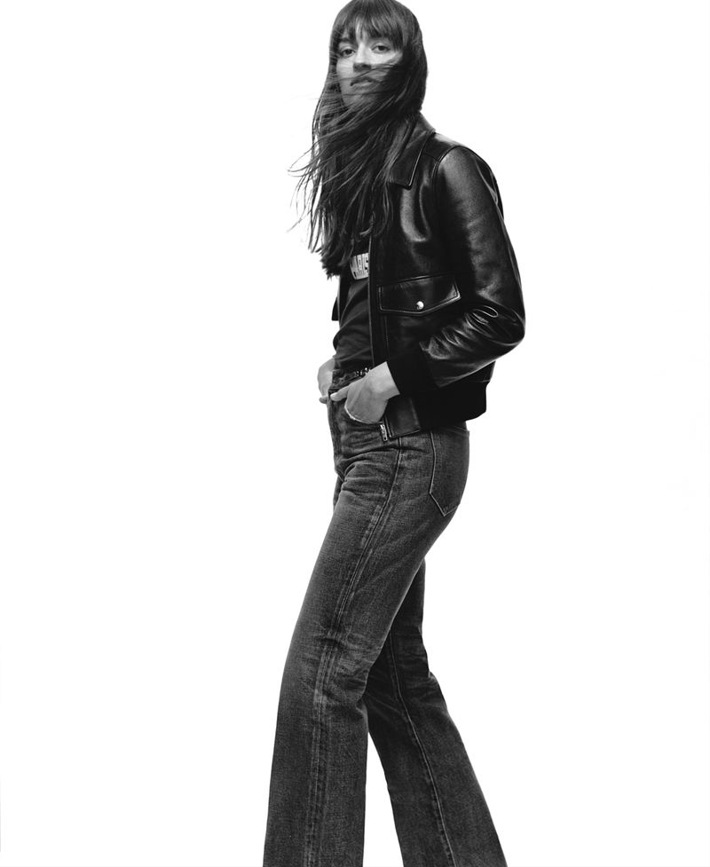 Black and white portrait of Clara Luciani posing in a leather jacket and wide-leg denim jeans against a plain white studio backdrop.