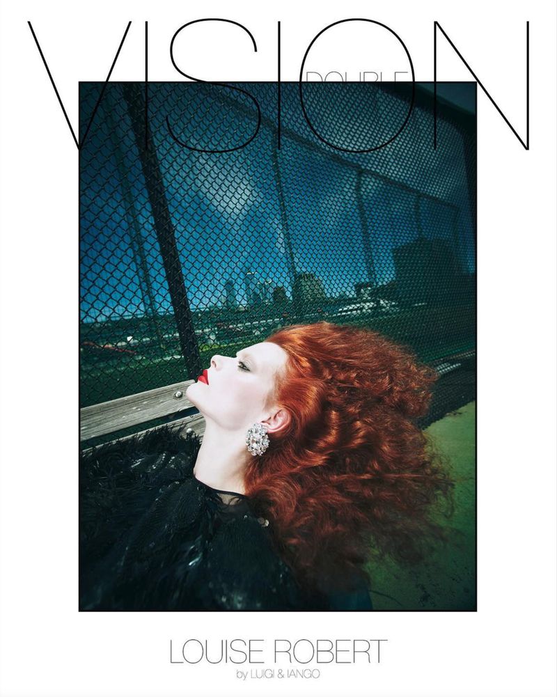 Model Louise Robert with voluminous red hair looking upward wearing a statement crystal earring and black attire against a chain-link fence.