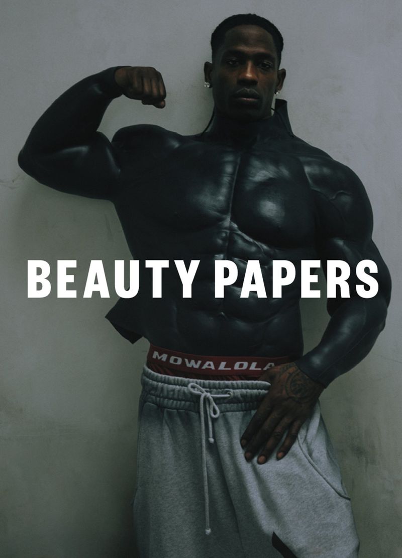 A muscular Black man poses in gray sweatpants and a faux muscle shirt in front of a light gray wall, promoting Beauty Papers.