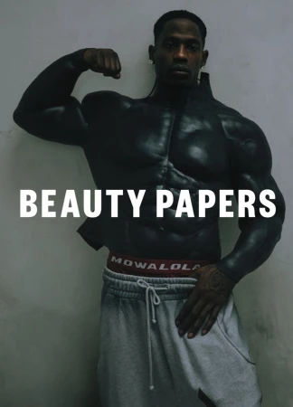A muscular Black man poses in gray sweatpants and a faux muscle shirt in front of a light gray wall, promoting Beauty Papers.