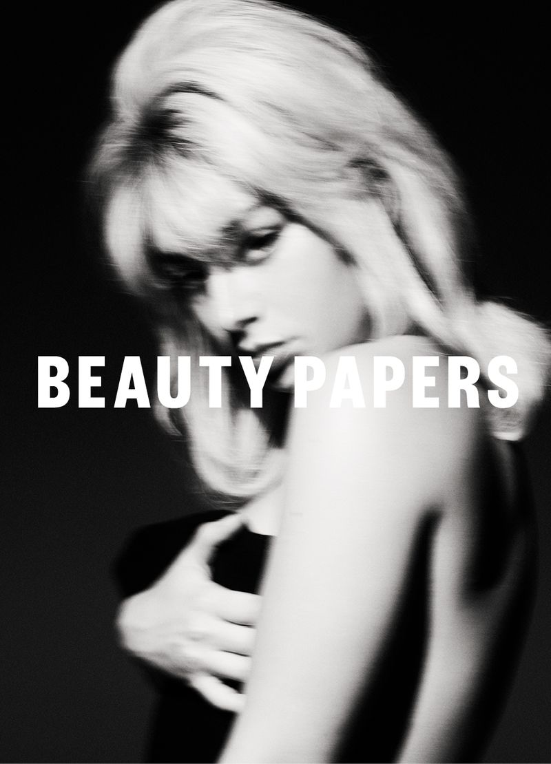 Black-and-white portrait of a young woman with blonde hair, partially nude, covering her chest with her arm, with “BEAUTY PAPERS” text overlay.