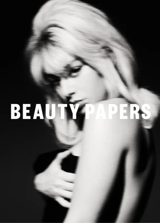 Black-and-white portrait of a young woman with blonde hair, partially nude, covering her chest with her arm, with “BEAUTY PAPERS” text overlay.