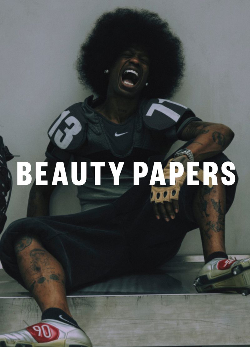 An athletic man with an afro in a football jersey and soccer shoes sitting against a gray wall, the words 'Beauty Papers' on the image.