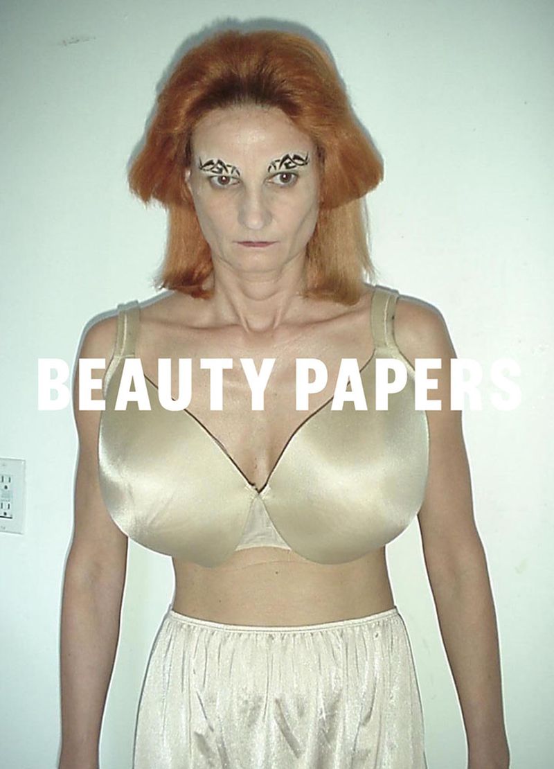 A mature woman with copper-colored hair and dramatic eye makeup, wearing vintage lingerie, stands in front of a neutral white wall.