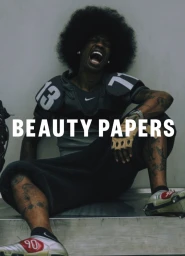Man with large afro hair wearing football gear and cleats laughing on a grey background