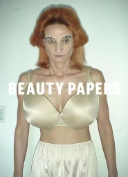 Model with red hair and brow designs wearing a beige bra against a white wall for Beauty Papers.