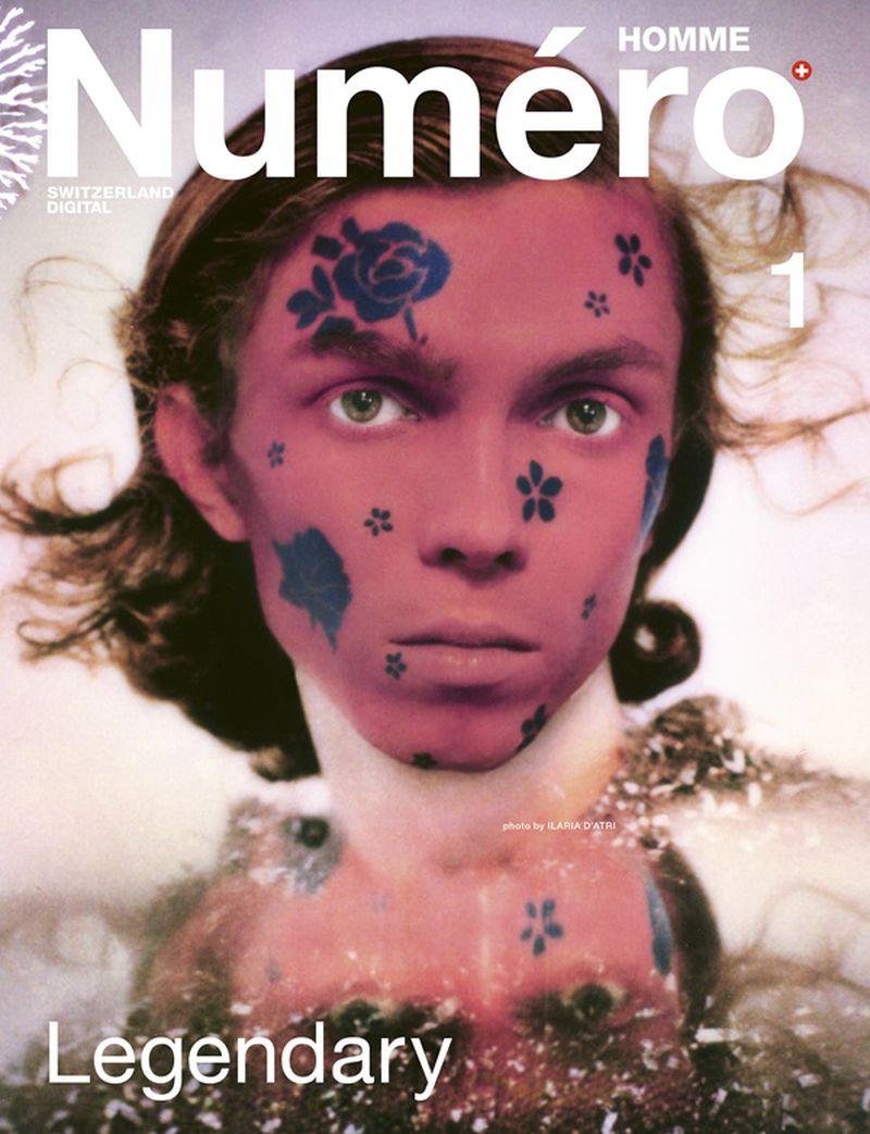 Close-up portrait of a model with blue floral face tattoos and reddish skin tone against a light background for Numéro Homme Switzerland