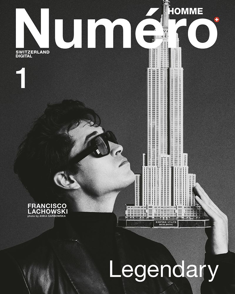 Man in sunglasses holds Empire State Building model on Numero Homme magazine cover, wearing black turtleneck and leather jacket in studio shot.