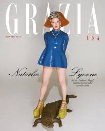 Natasha Lyonne wearing a blue dress and platform shoes standing on a bronze crocodile sculpture against a white backdrop.