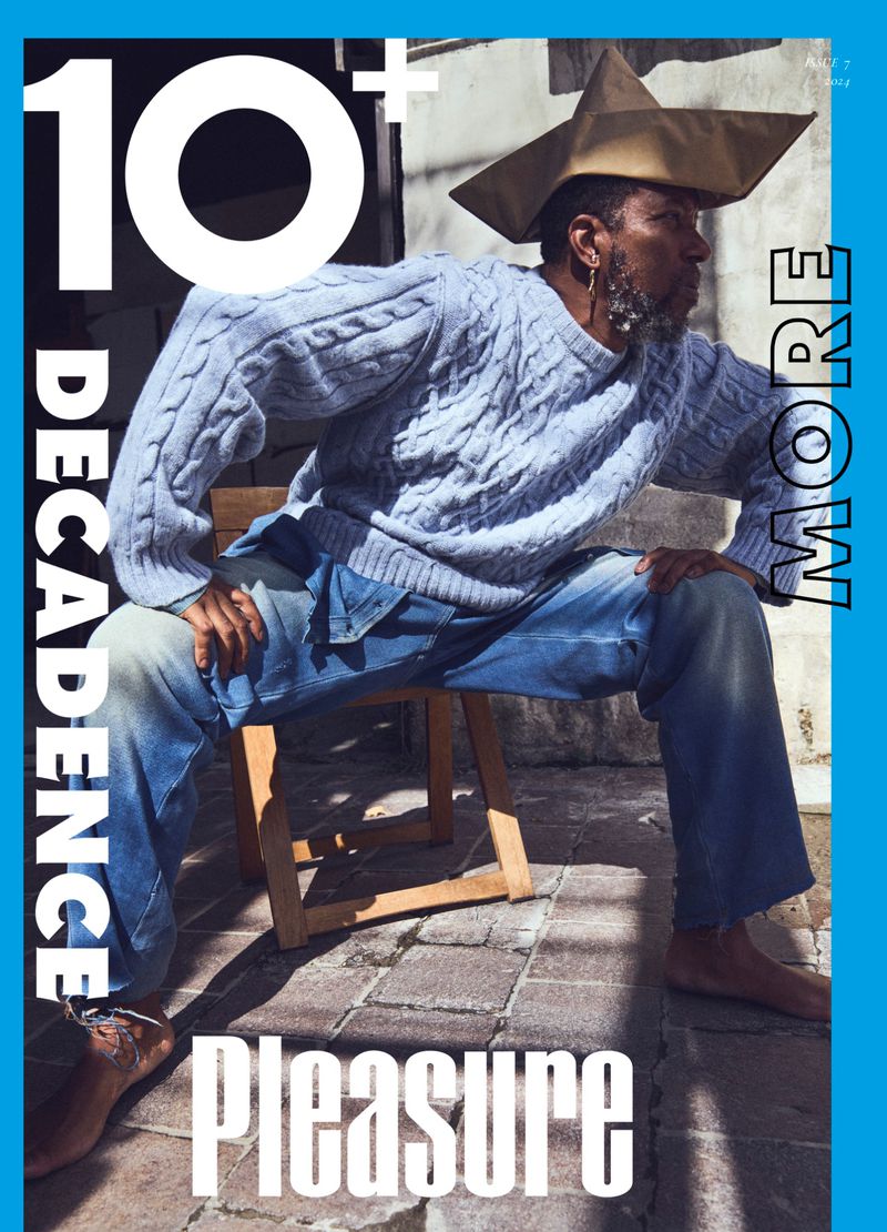 Magazine cover featuring a stylish adult male in a paper hat and cable-knit sweater, seated outdoors against a neutral backdrop, with bold text.