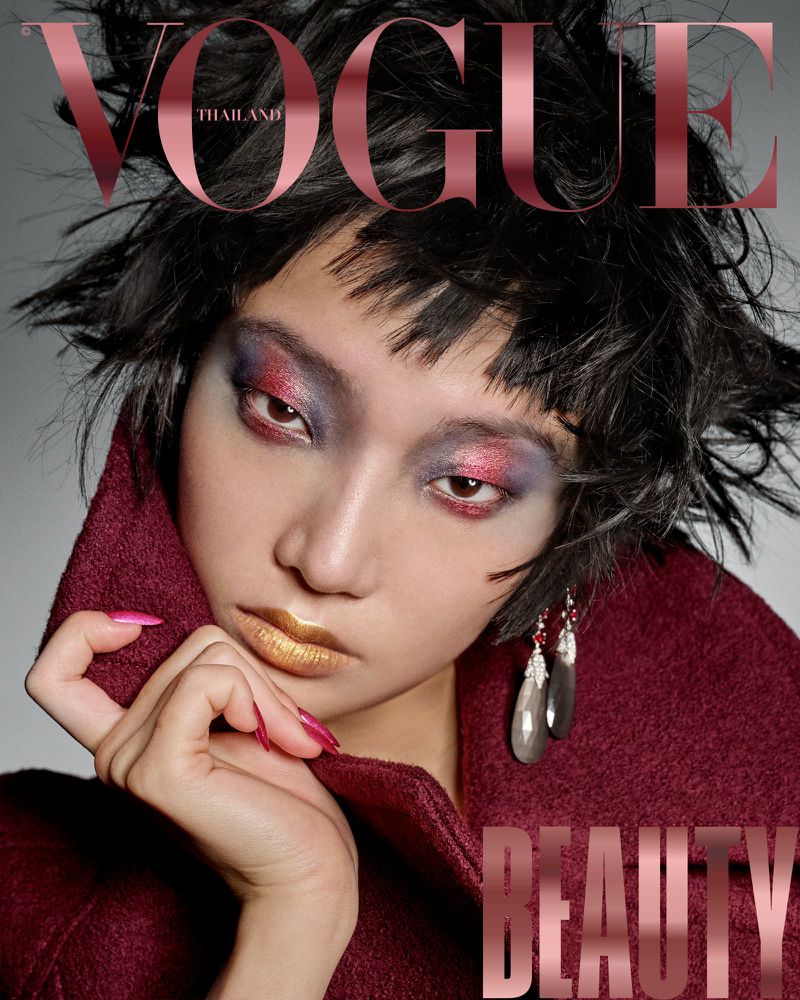 Vogue Thailand magazine cover features a young woman with bold makeup, wearing a burgundy coat, and silver earrings on a gray background.