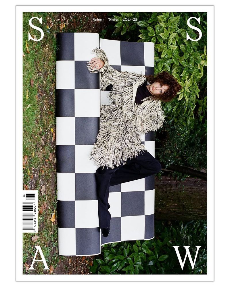 Magazine cover featuring a woman in a shaggy jacket reclining on a black and white checkered surface in a lush, green outdoor setting.