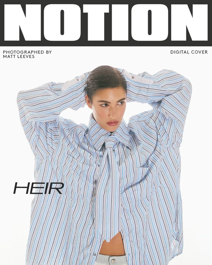 Model with slicked-back hair wearing an oversized blue striped shirt with hands behind head against a white background