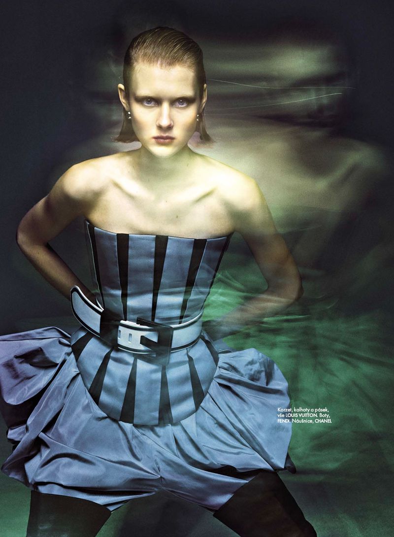 Fashion editorial image featuring a woman in a blue corset dress with a black belt, posing against a blurred, ethereal green backdrop.