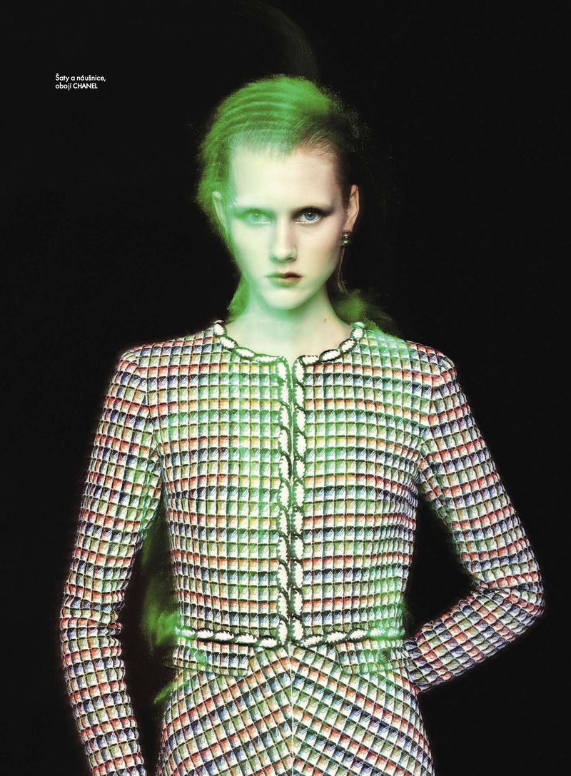 A young woman with light skin poses in a fitted tweed jacket and skirt ensemble, illuminated by green light, set against a black background.