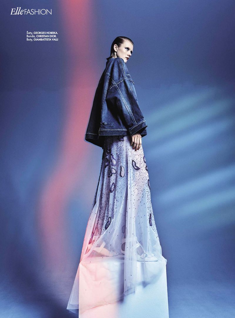 Fashion editorial shot of a woman in a denim jacket and embellished gown, posing on a white cube against a blue backdrop with soft lighting.