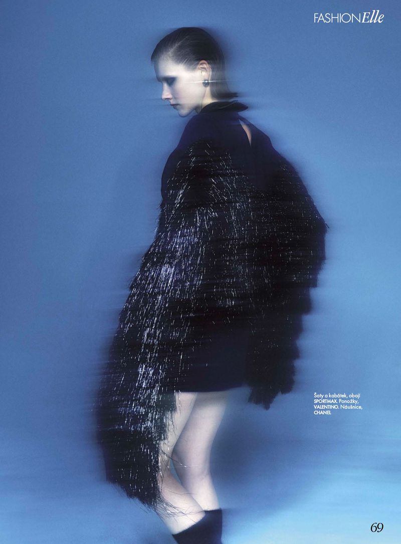 Fashion editorial shot of a slender young woman in a black dress with fringed shawl against a blurred light blue background.